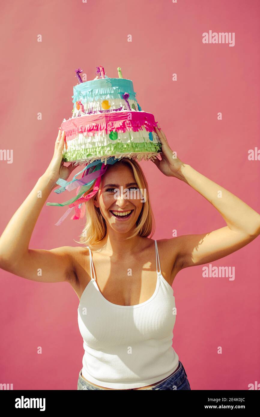 Cheerful woman carrying birthday cake on head against pink wall Stock