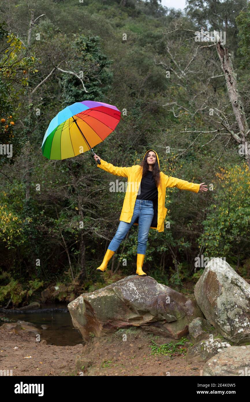 Carefree young woman wearing yellow raincoat holding umbrella while standing on rock in forest Stock Photo