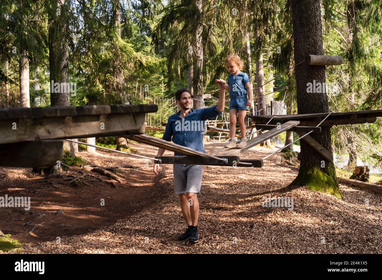 Father assisting little daughter walking across small suspension bridge on forest obstacle course Stock Photo