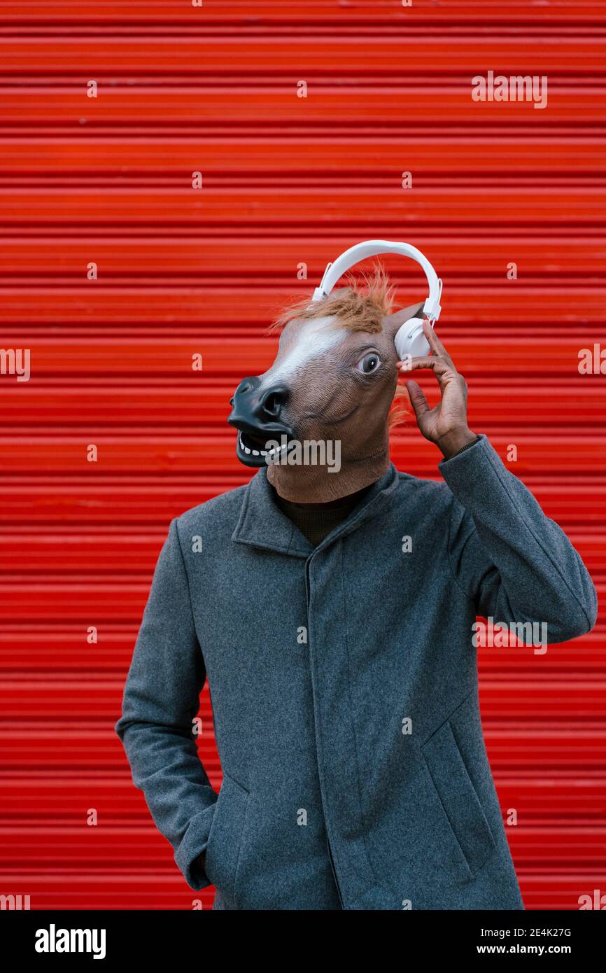 Man with horse mask wearing headphones while standing against red wall