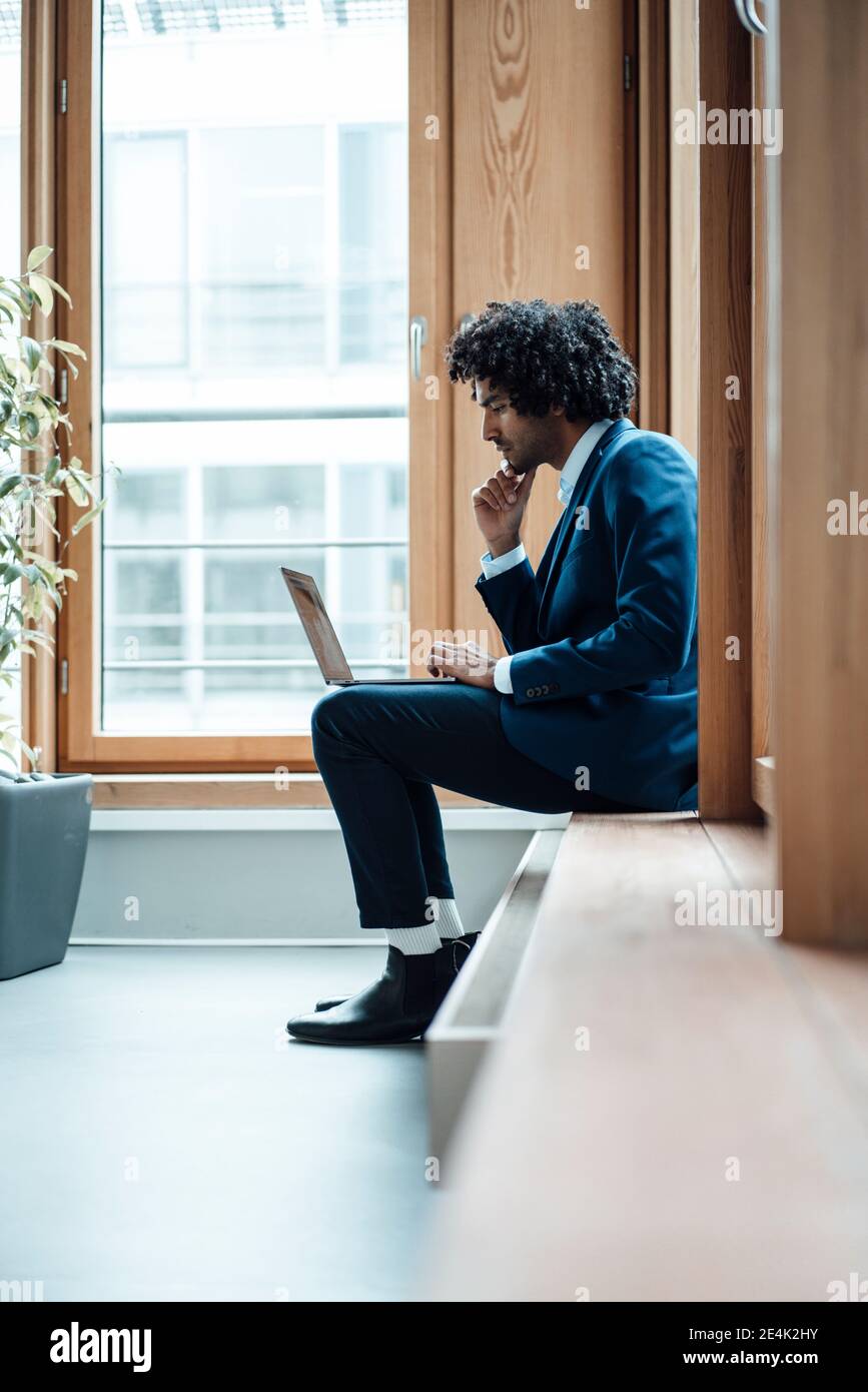 Young businessman using laptop while sitting against window at workplace Stock Photo