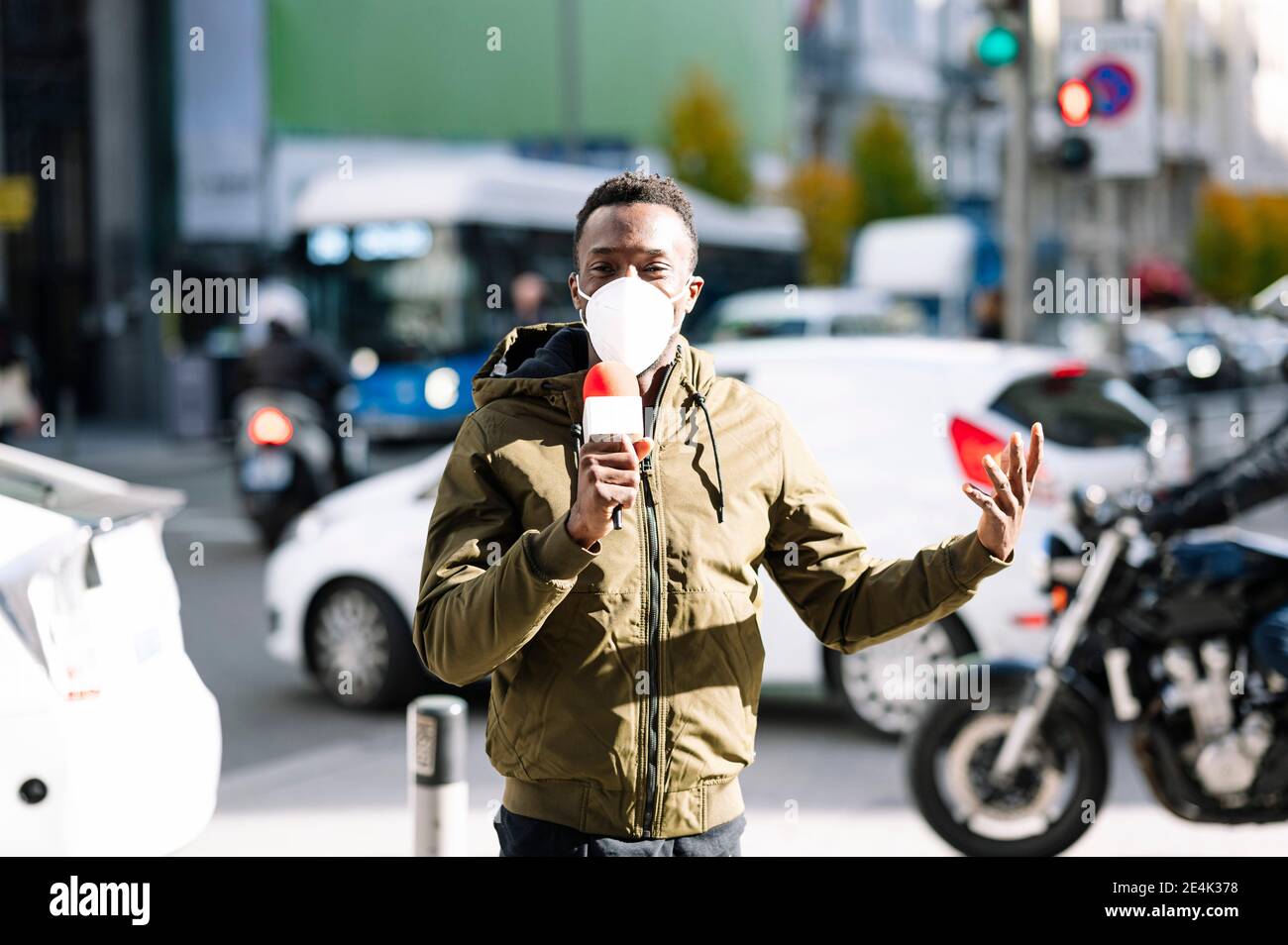 Male TV reporter wearing face mask talking over microphone while standing on street Stock Photo