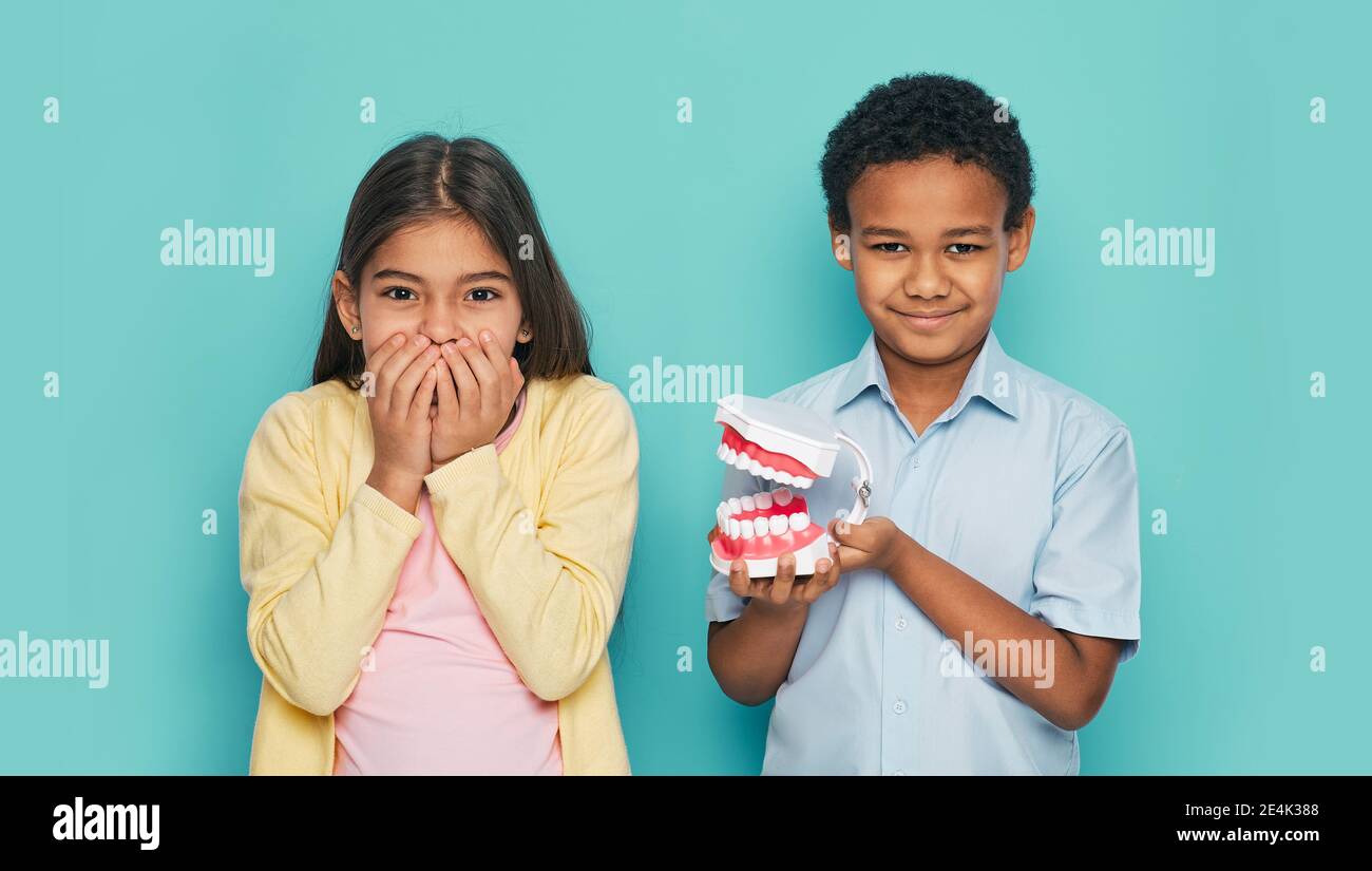 Scared multi-ethnic children showing toothache on a turquoise background. Girl hiding her mouth behind hands and boy holding jaw model in his hands Stock Photo