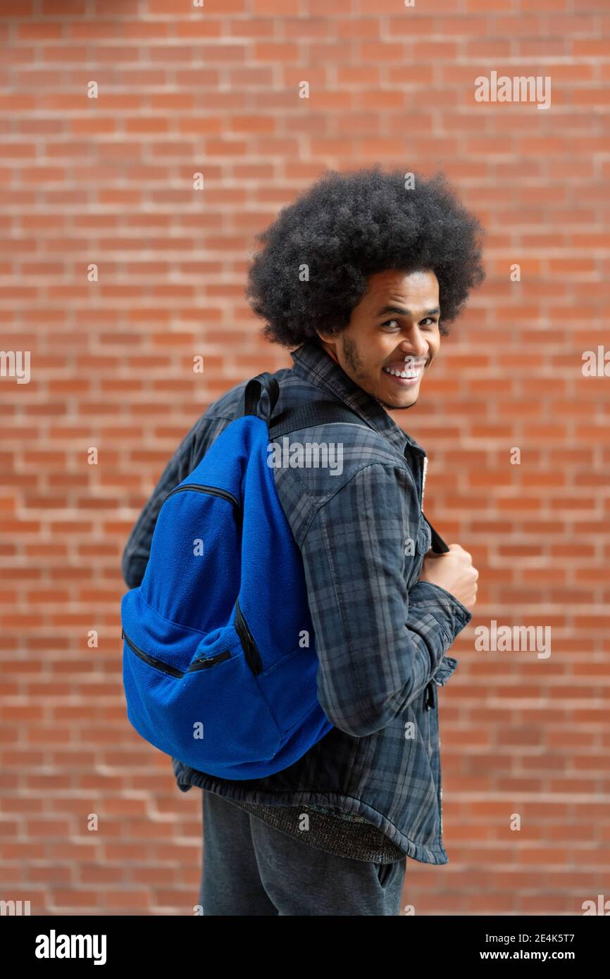 Cheerful young man with blue backpack standing in front of brick wall Stock Photo