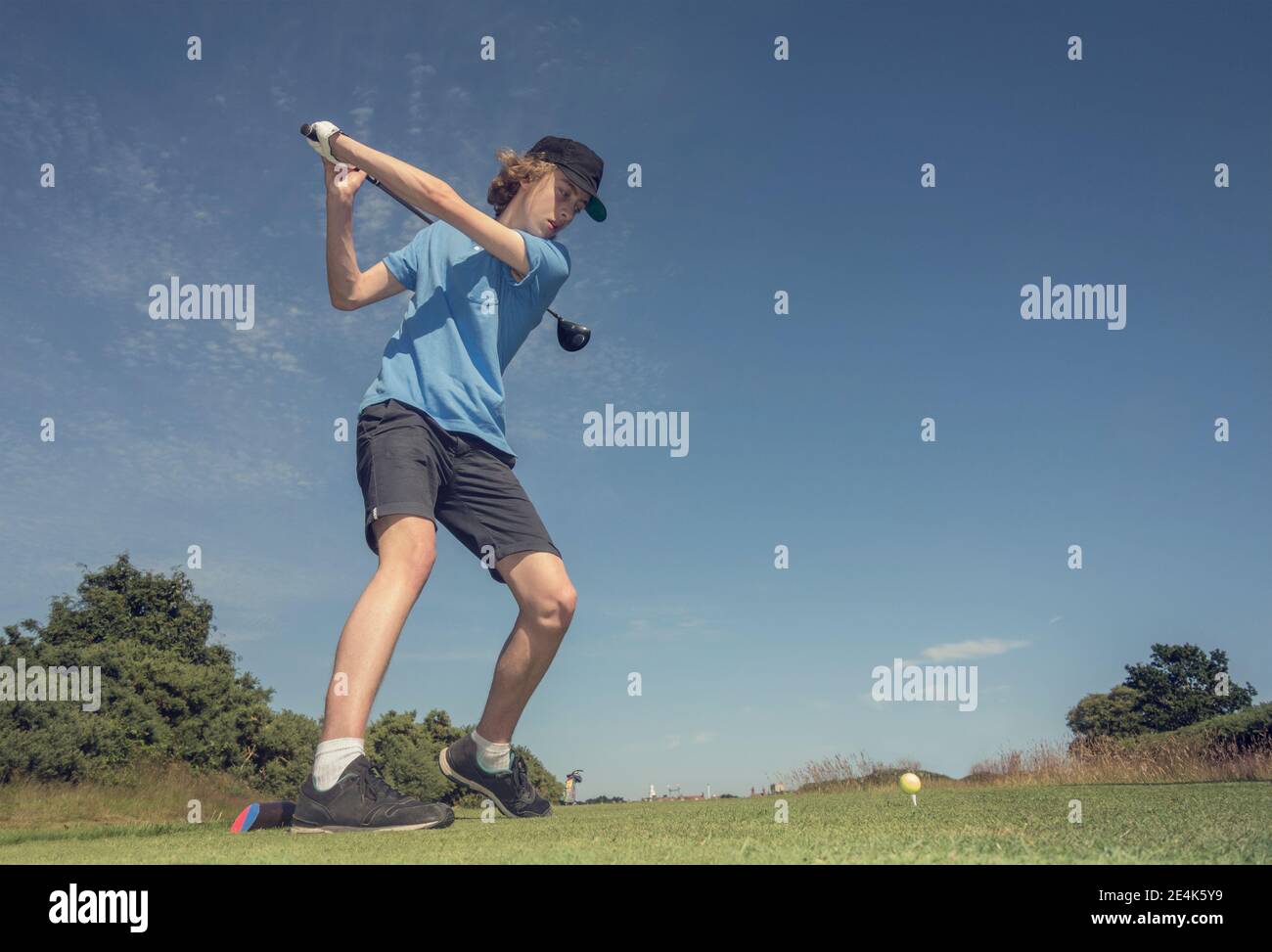 Teenage boy hitting golf shot against blue sky at course Stock Photo