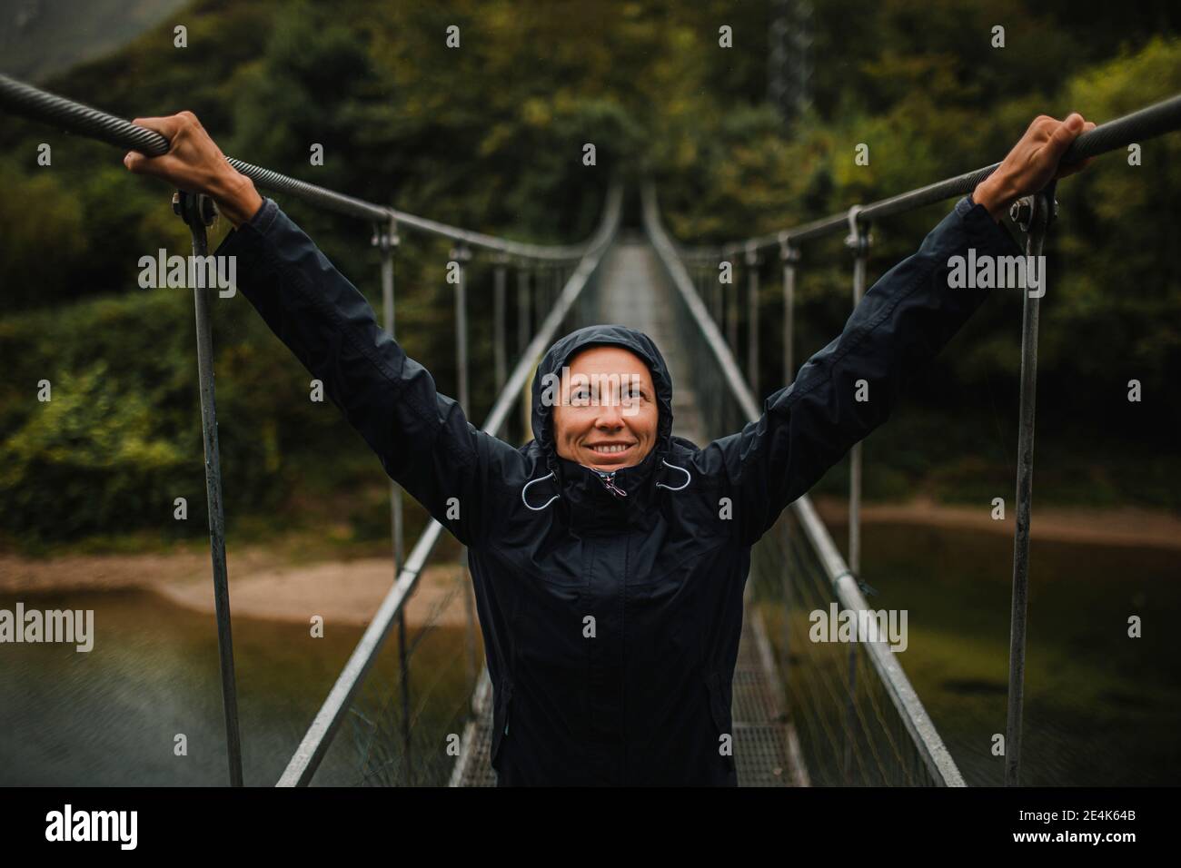 Smiling woman wearing raincoat with arms raised standing on suspension bridge over Sella river in forest Stock Photo