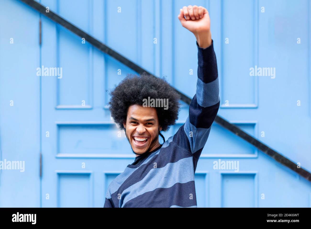 Cheerful young man with arm raised screaming while standing against blue wall Stock Photo