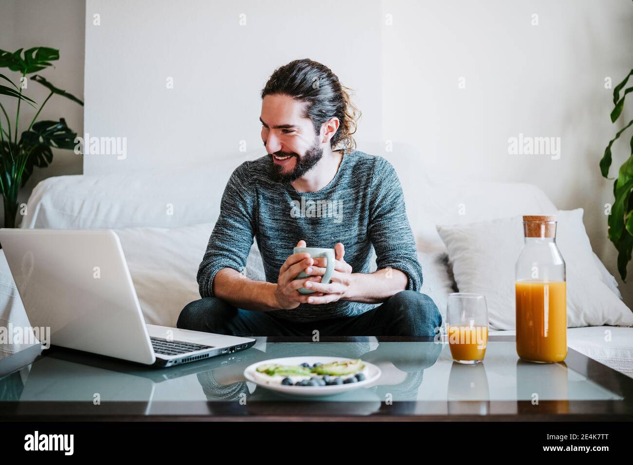 Young man with coffee cup using laptop while having breakfast sitting on sofa at home Stock Photo
