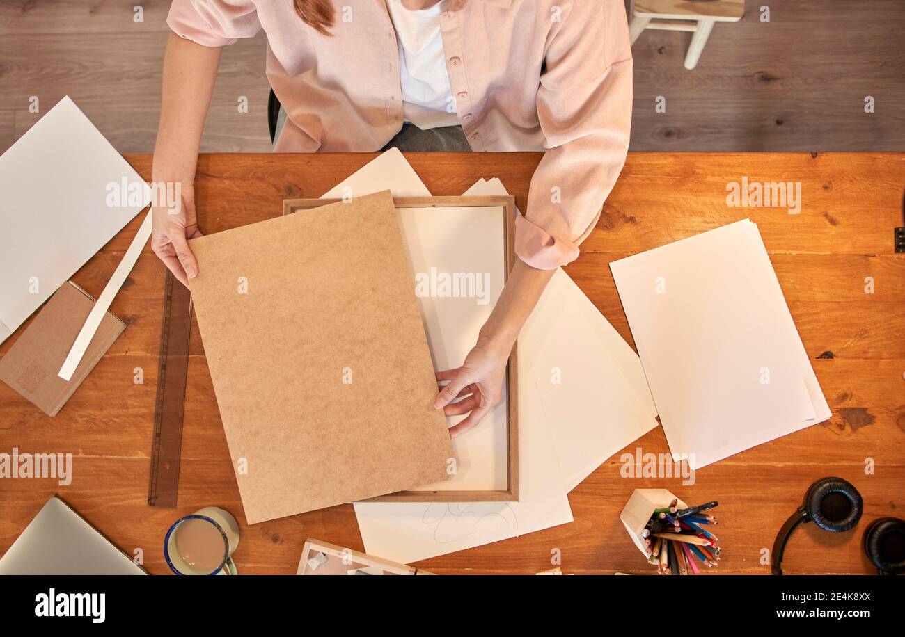 Female artist holding wooden picture frame at desk while working at home Stock Photo