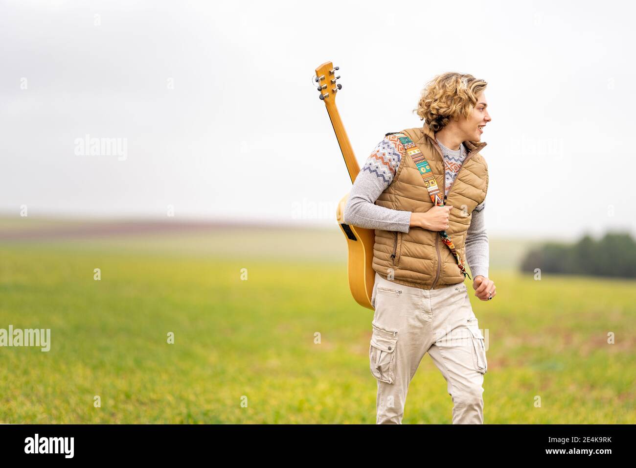 Young man walking across grassy field with acoustic guitar on back