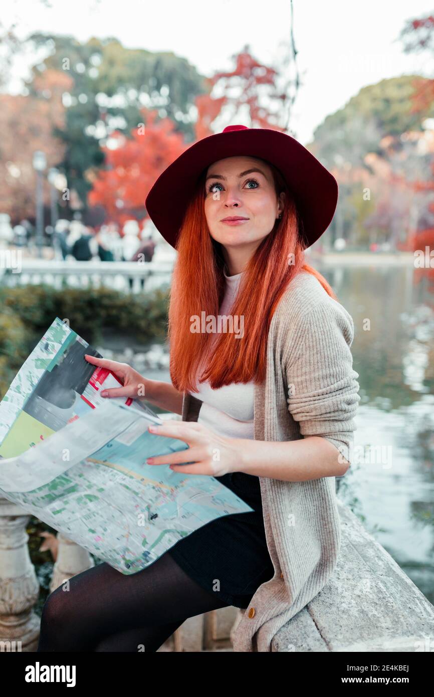 Beautiful woman wearing hat holding map while sitting in park Stock Photo