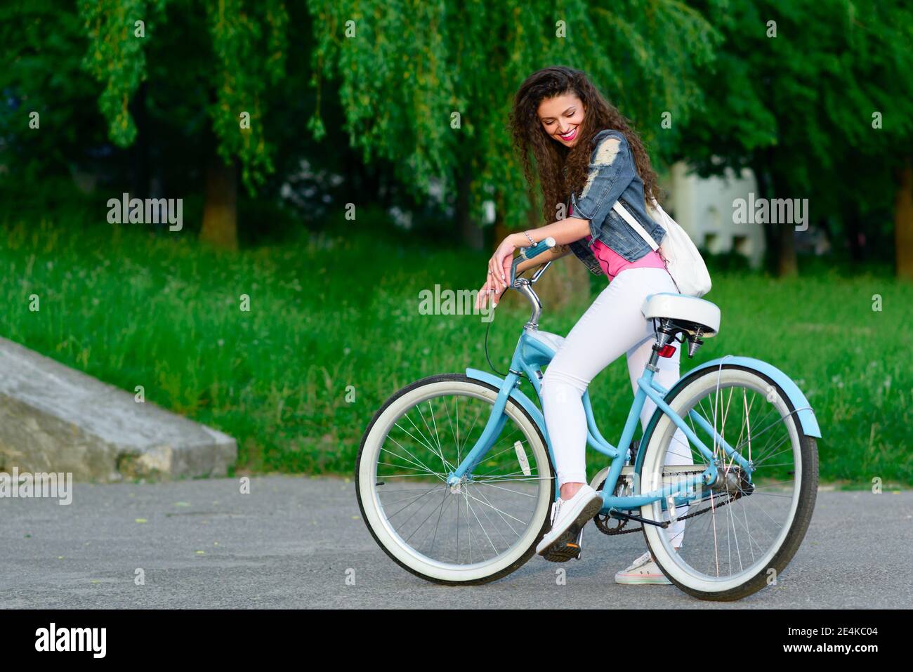 Young girl in casual clothes with a beautiful smile and curly hair rides a bicycle in the city in the summer Stock Photo