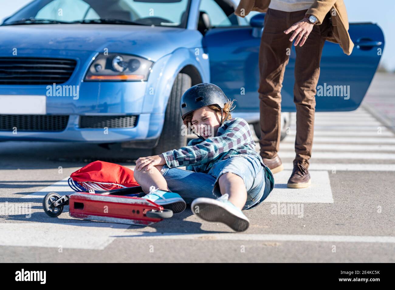 Boy falling down from push scooter with man standing by car on road after accident Stock Photo