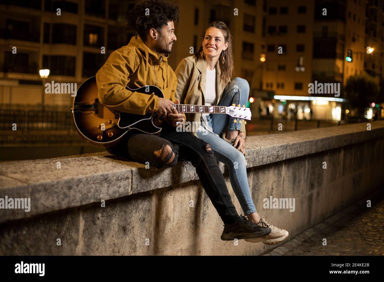 Couple with guitar smiling while sitting on retaining wall Stock Photo