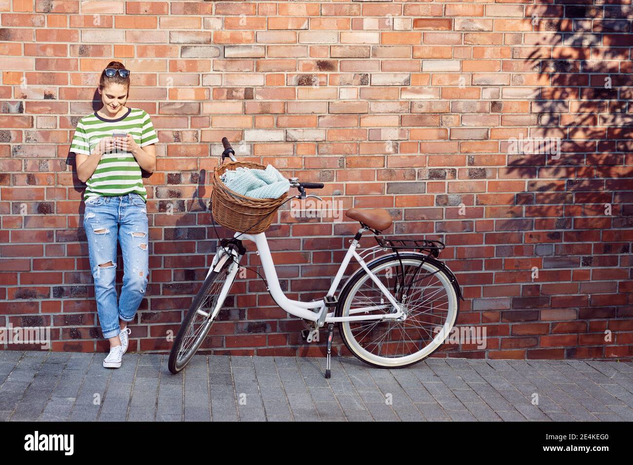 Woman text messaging through mobile phone while bicycle parked on footpath against brick wall Stock Photo