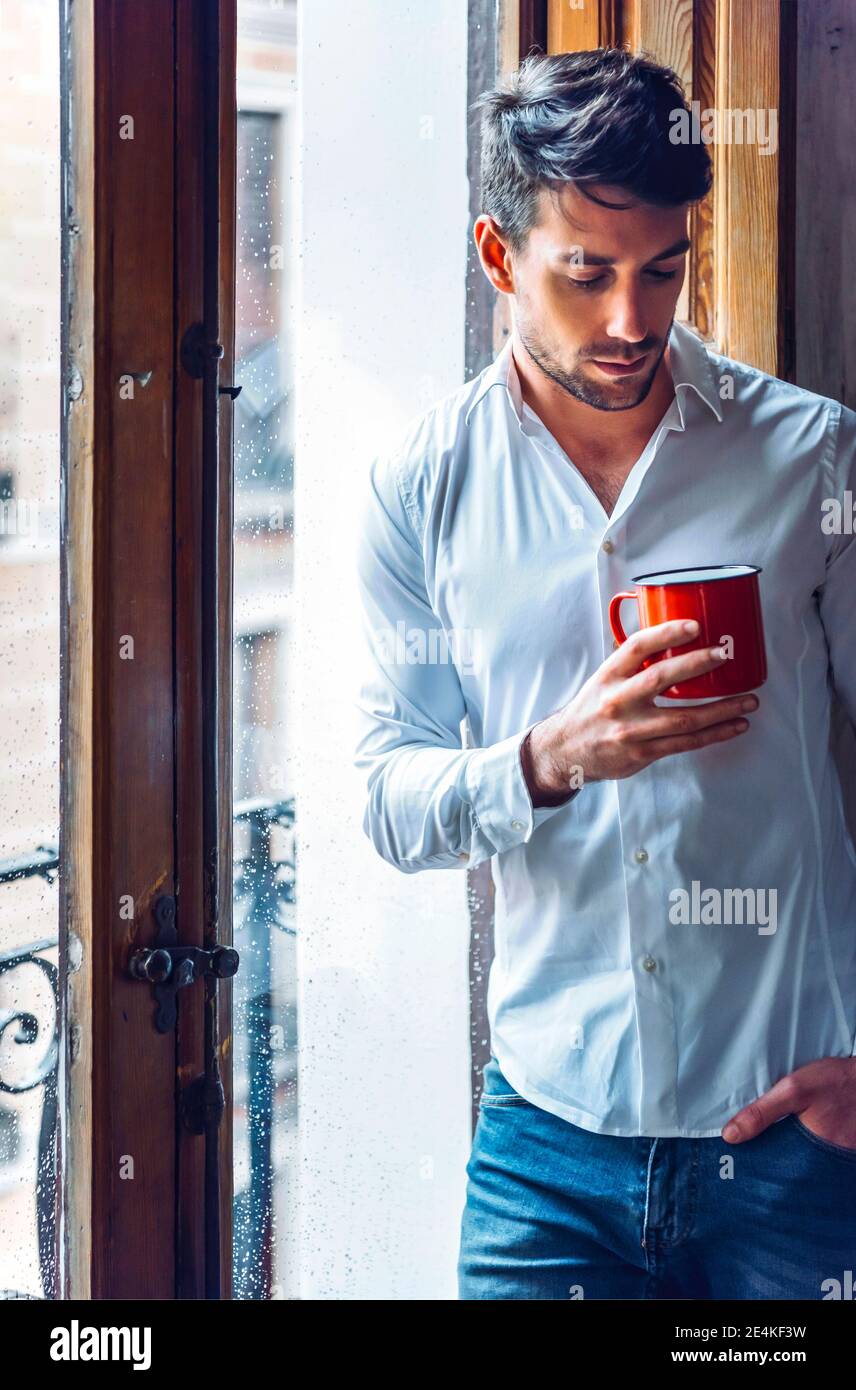 Thoughtful young man holding cup standing at the window Stock Photo