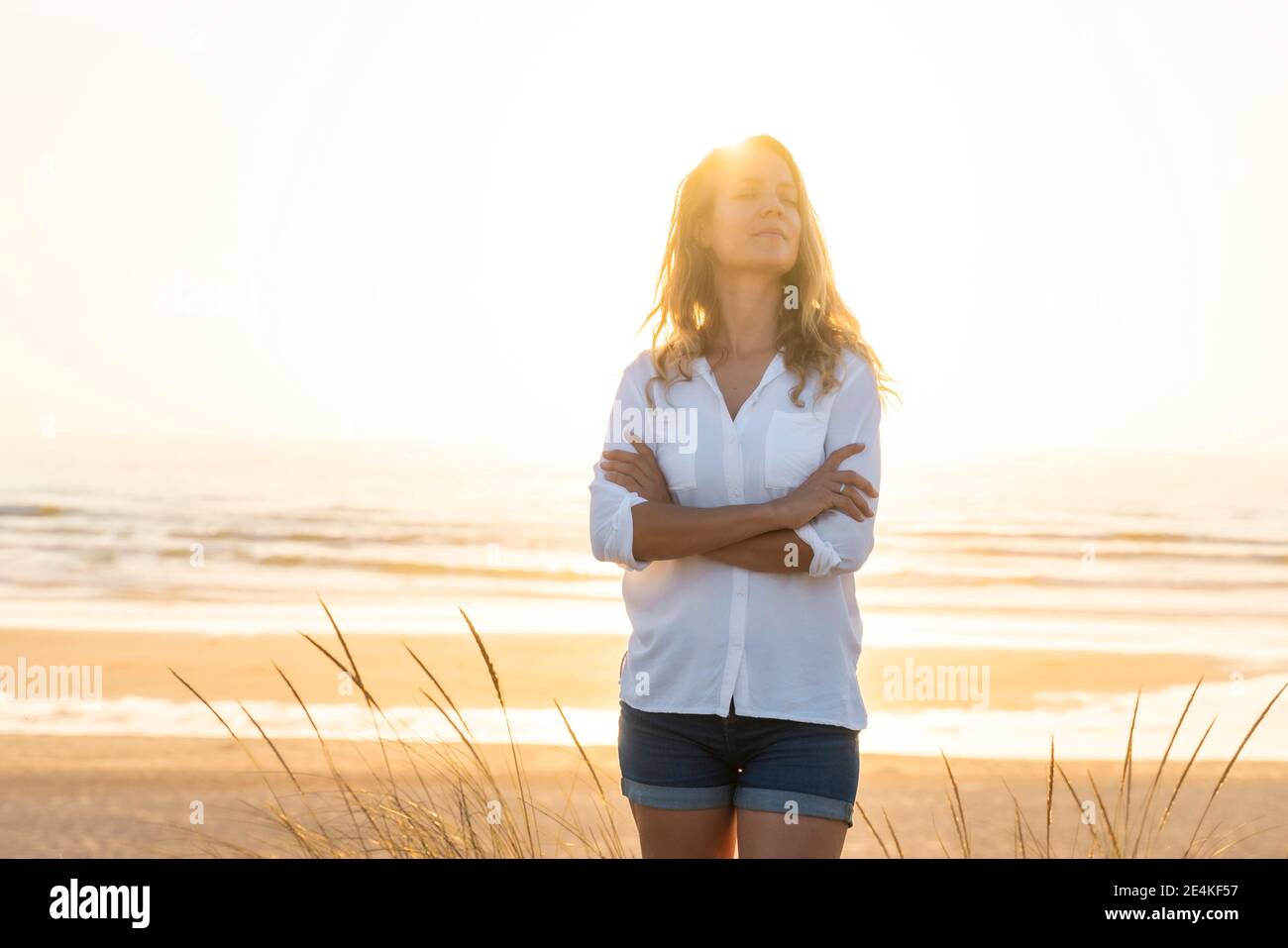 Mid adult woman standing wit arms crossed at beach during sunset Stock Photo