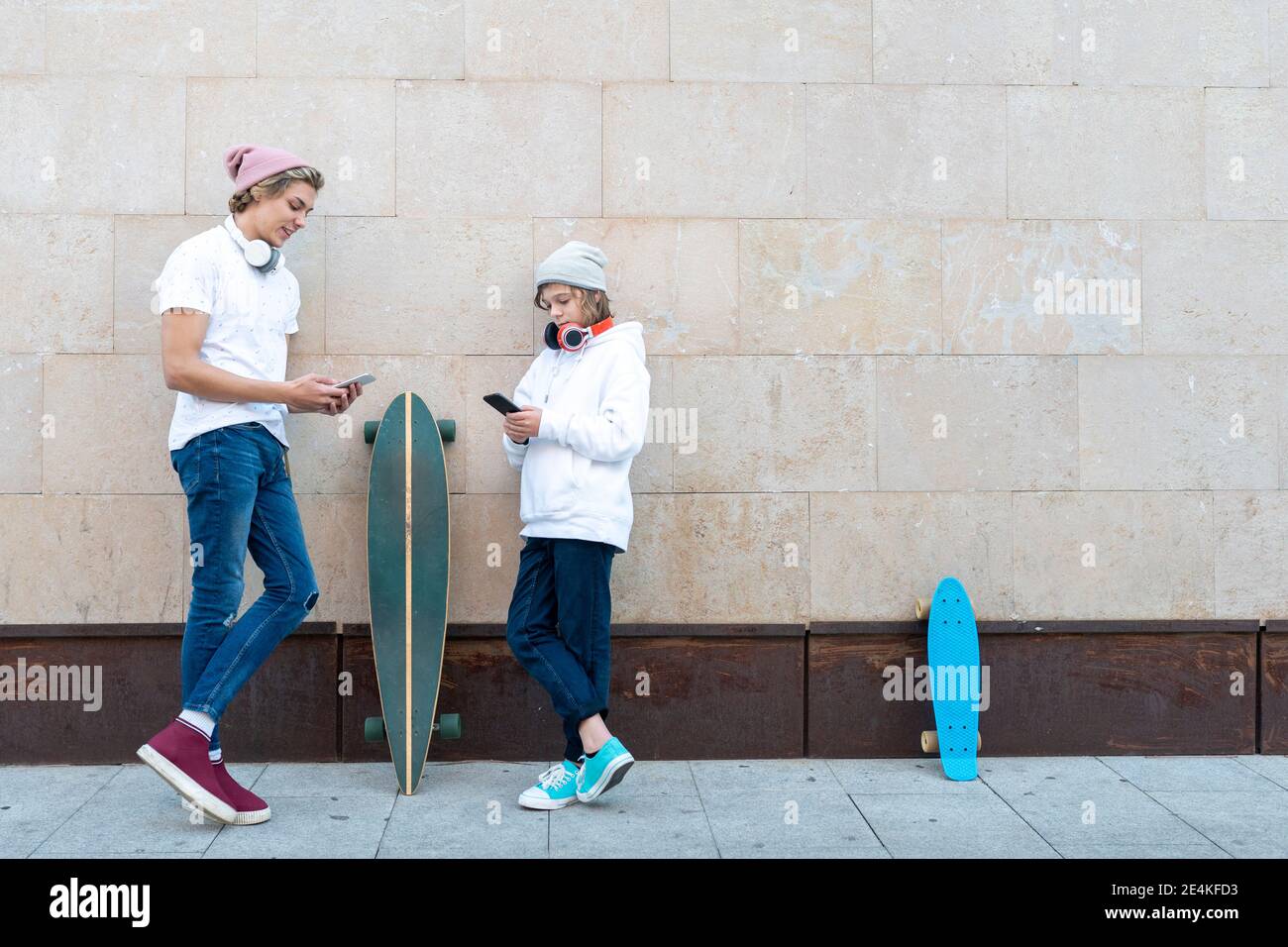 Young man and boy wearing knit hat using mobile phone while standing by skateboard against wall Stock Photo