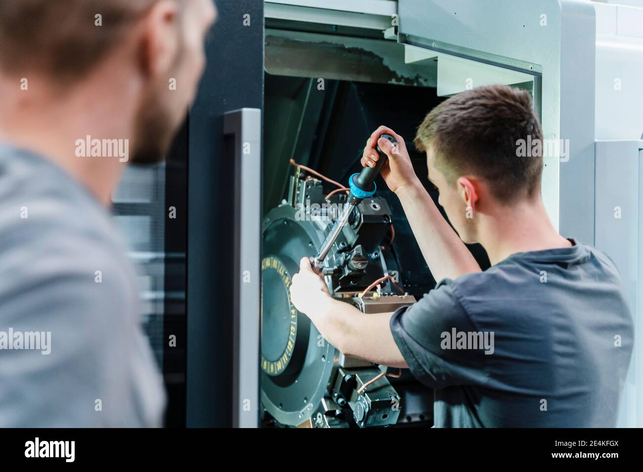 Male trainee fastening bolts of machine while standing at industry Stock Photo