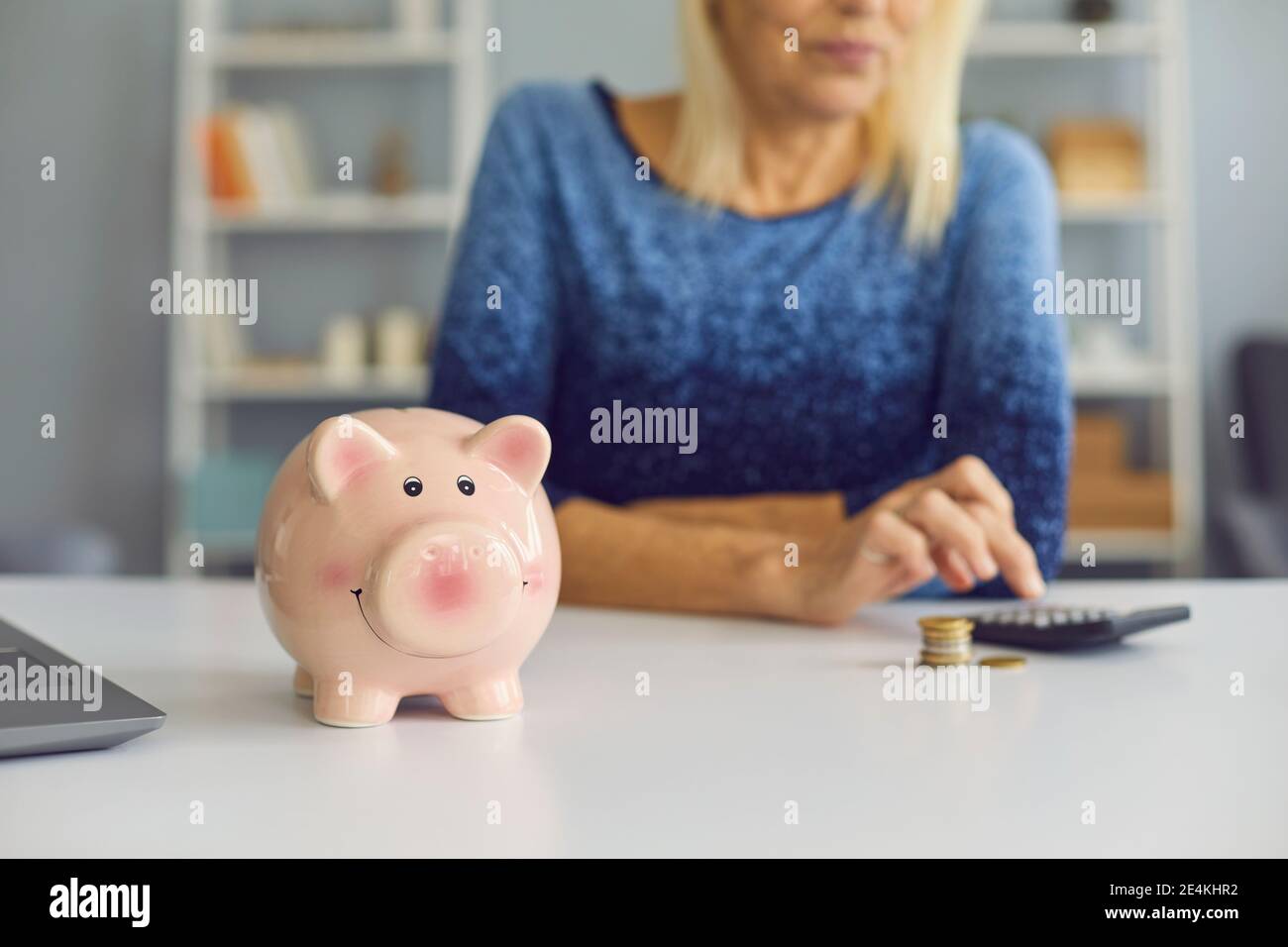 Mature woman sitting at table with piggy bank, using calculator and ...
