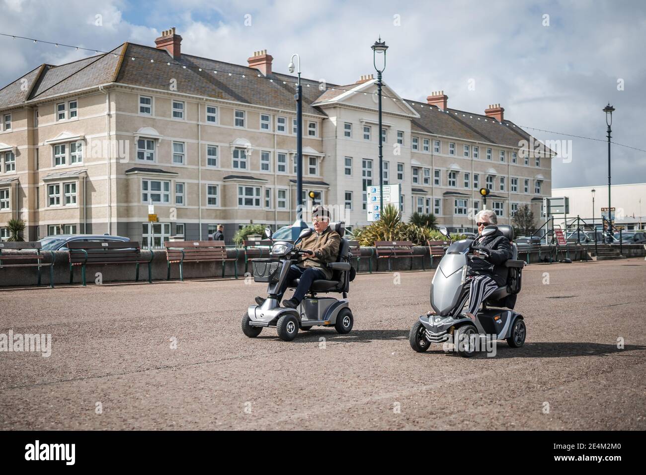 Two pensioners on mobility scooters riding along sea front seaside hats on cruising along racing race llandudno cool old people escaping folks home Stock Photo