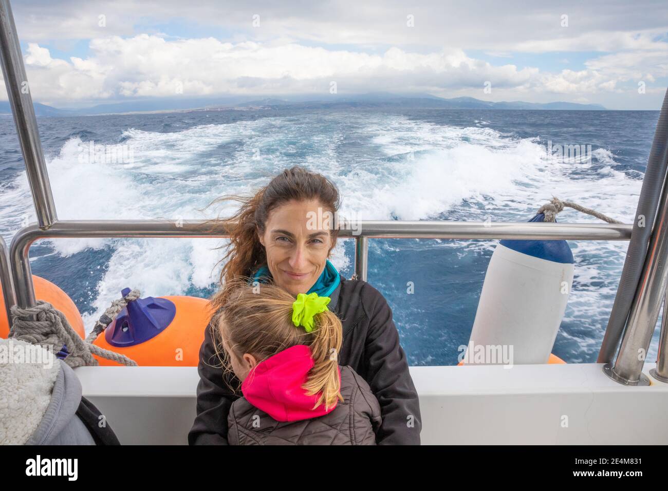 mother, smiling and looking at, embracing her daughter, four years old blonde girl with pigtail, sitting on the stern of motorboat sailing from Tarifa Stock Photo