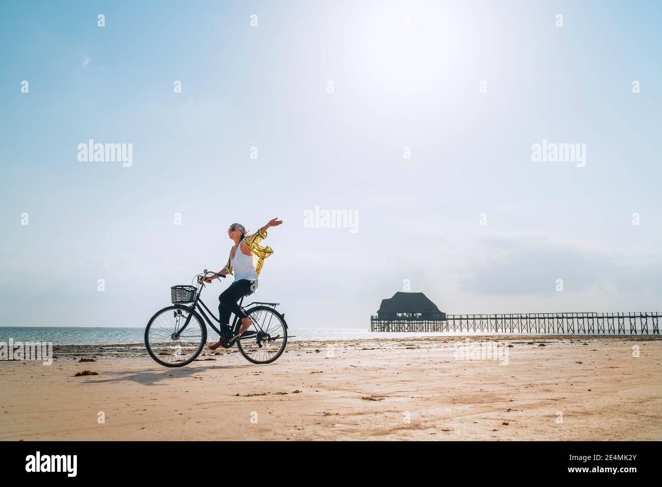 Young female dressed light summer clothes joyfully threw up her hand riding old vintage bicycle with front basket on the low tide ocean white sand coa Stock Photo