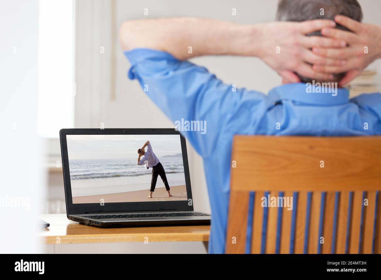 Businessman in home office doing stretching or relaxation exercise in front of laptop with man doing yoga on a beach - focus on the screen Stock Photo