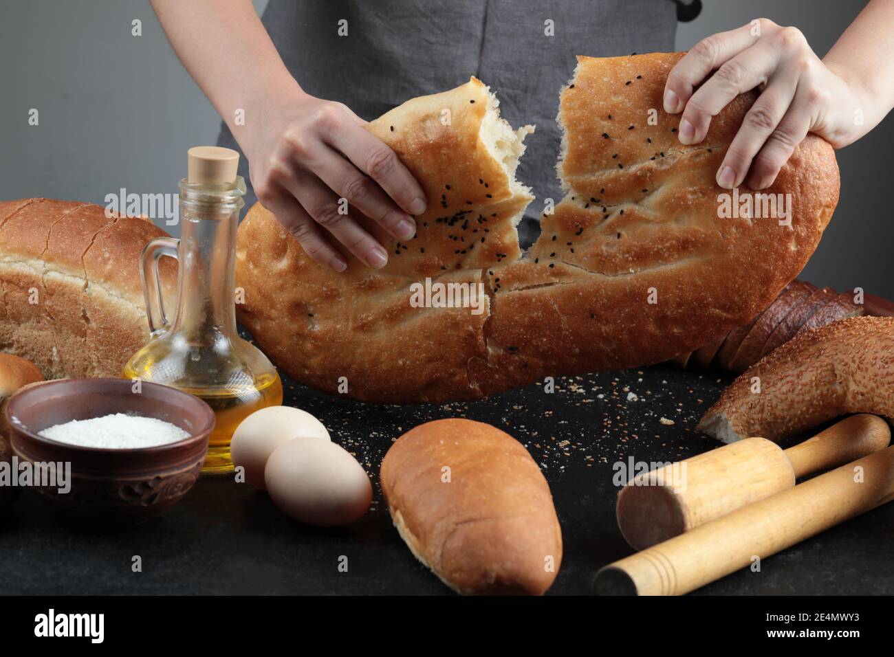 Woman cut bread into half on dark table with eggs, flour bowl and glass of oil Stock Photo