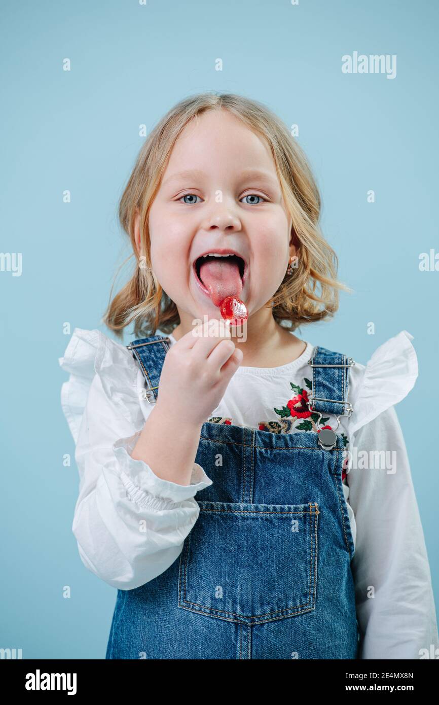 Happy little blonde girl enjoys eating lollipop candy over blue Stock Photo