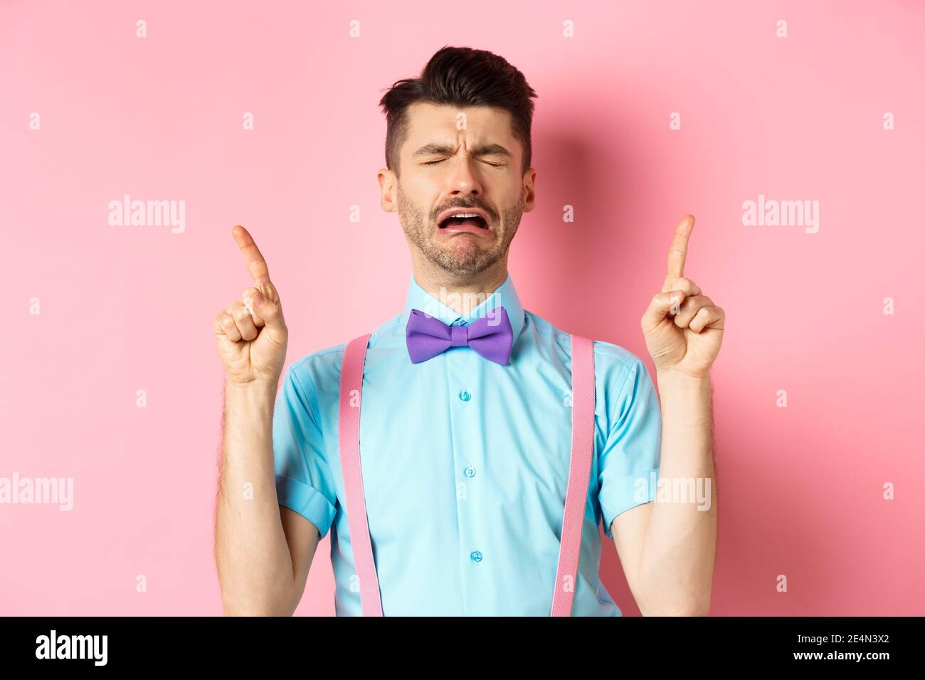 Sad and miserable guy sobbing and crying, pointing fingers up and something disappointed, standing upset on pink background Stock Photo