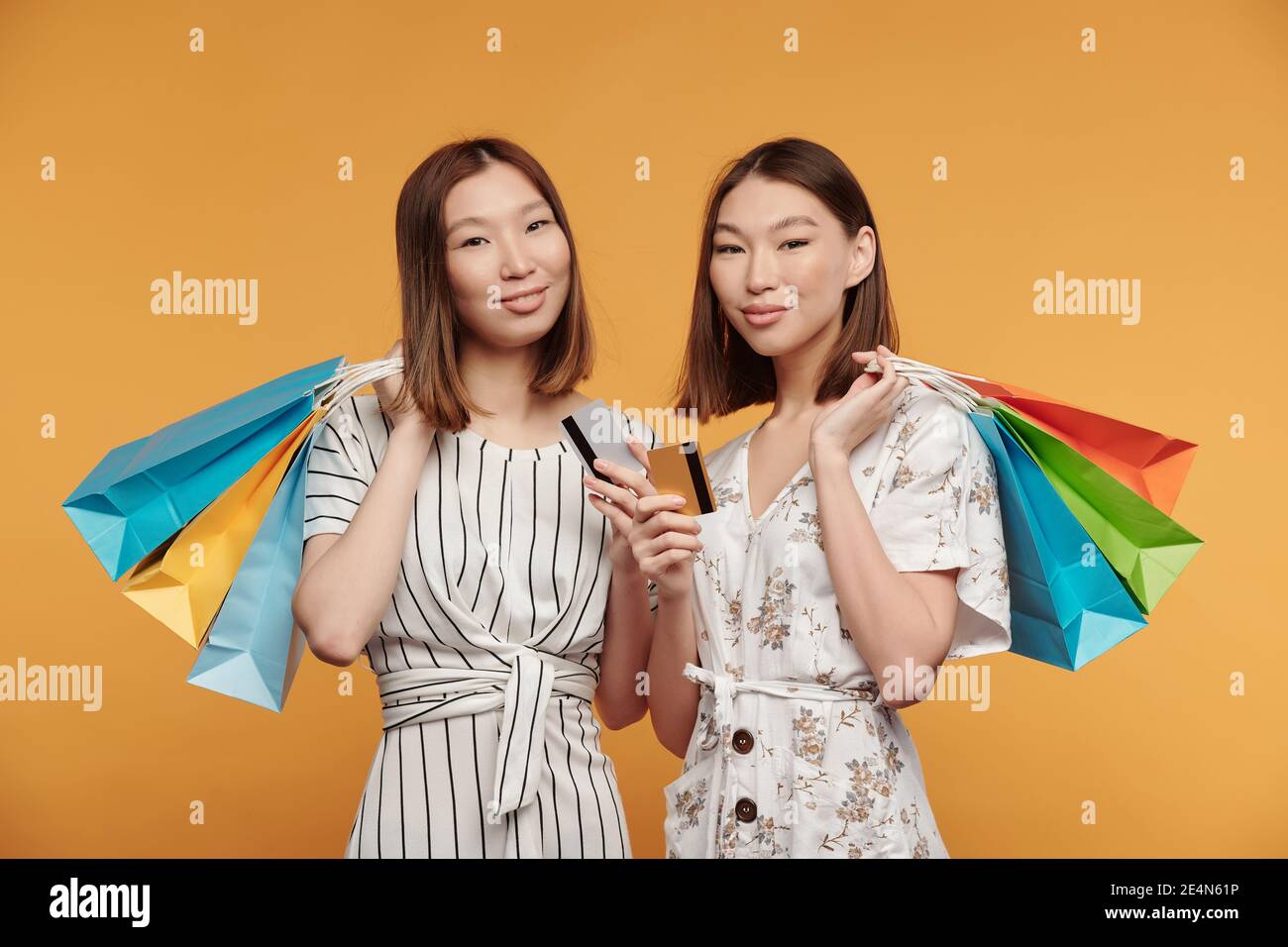 Two happy young Asian female twin consumers with paperbags and credit cards standing in front of camera against yellow background Stock Photo