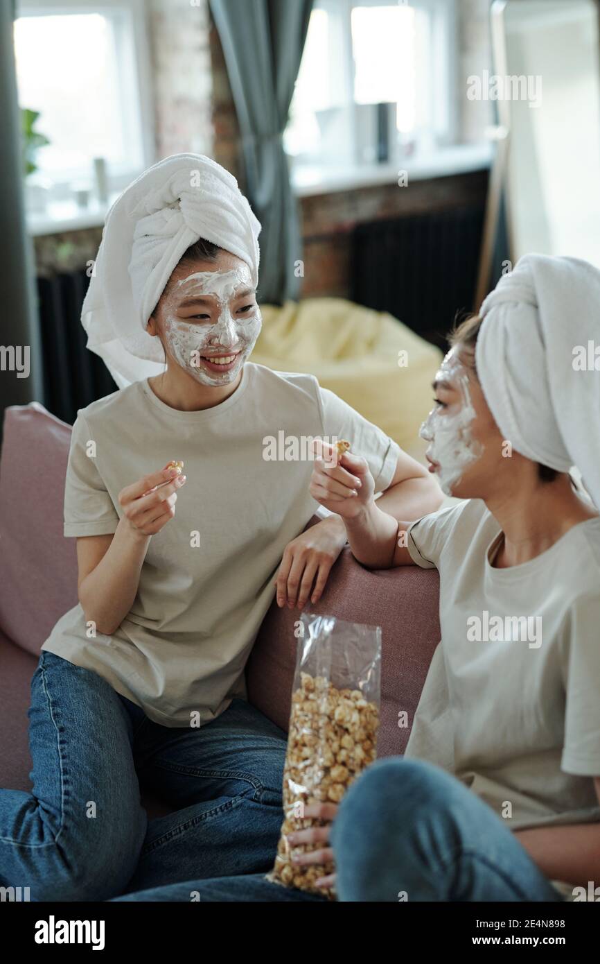 Young casual female twins with towels on heads and clay mask on faces sitting on couch at home, discussing curious stuff and having popcorn Stock Photo
