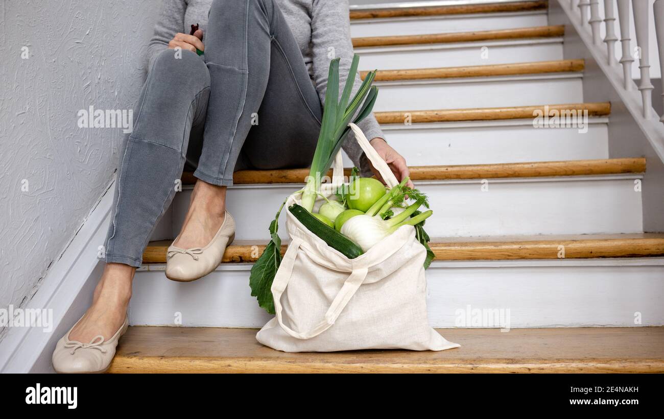 Woman with a plastic-free recycling groceries shopping bag and local  farm vegetables sits on the wooden staircase. Healthy vegan  sustainable life. Stock Photo