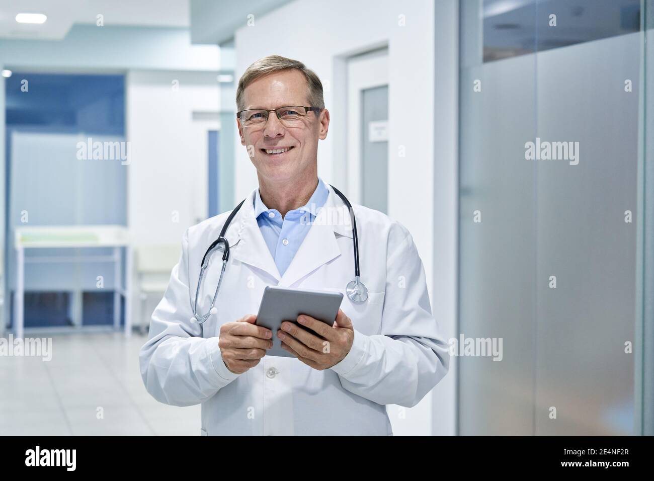 Smiling old middle aged doctor using digital tablet standing in hospital. Stock Photo