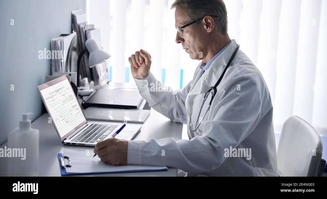 Serious older doctor using laptop computer in medical office. Stock Photo