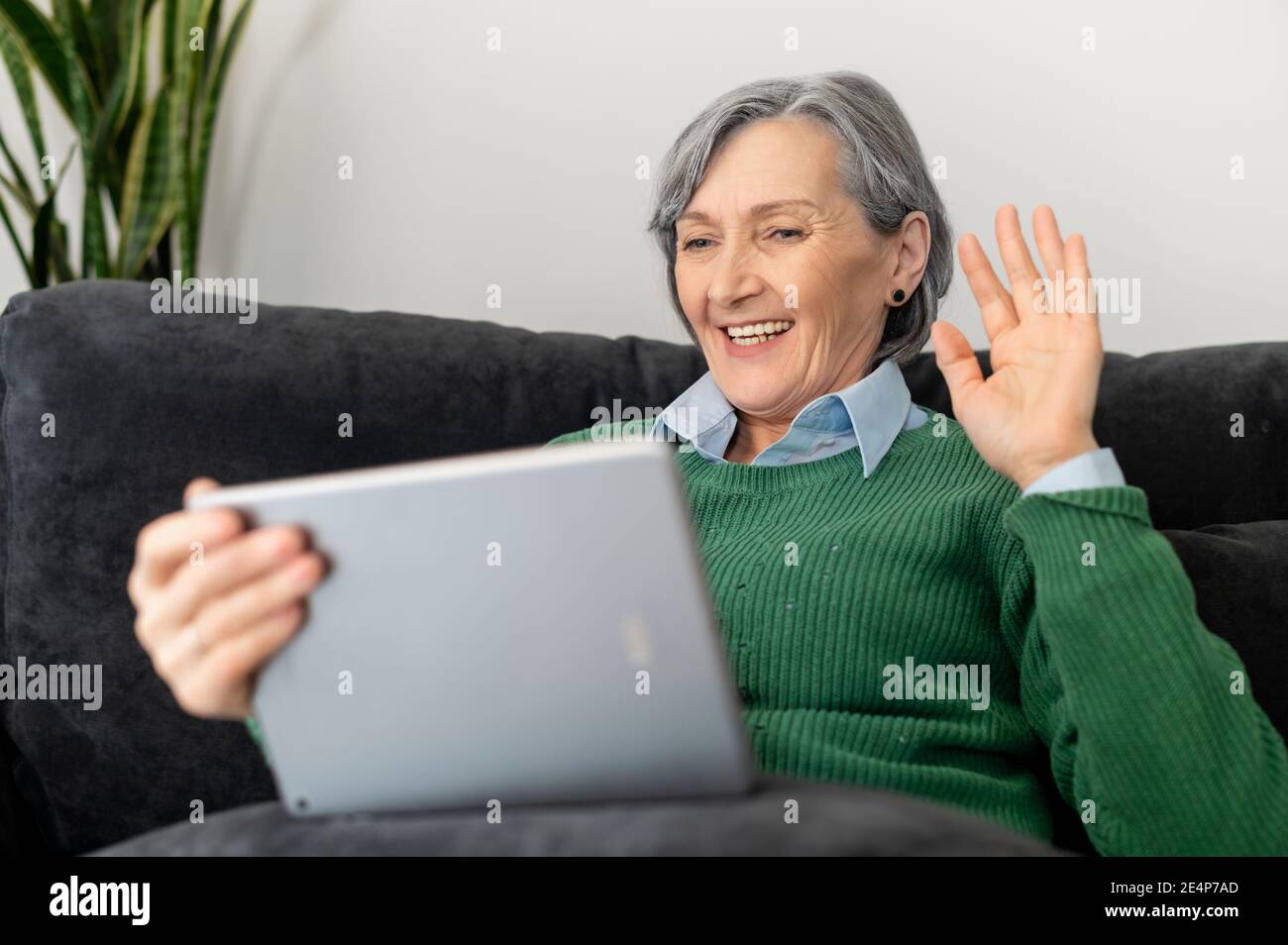 Positive senior mature lady in a green jumper talking online on the touchpad, watching a webinar, video-calling with the family and coworkers, waving Stock Photo