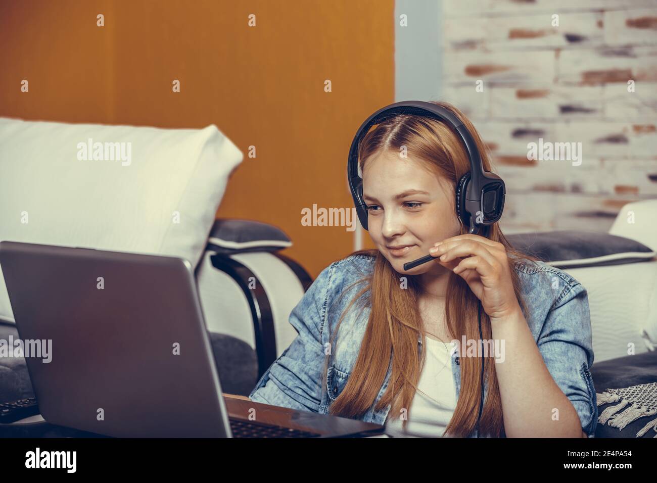 Happy teenage girl sitting on the floor wearing headphones, taking exam during a video call online with a laptop in the living room. Girl actively ans Stock Photo