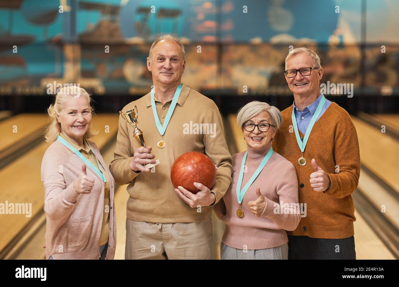 Waist up portrait of smiling senior team wearing medals holding trophy and looking at camera while standing in bowling alley after winning match Stock Photo