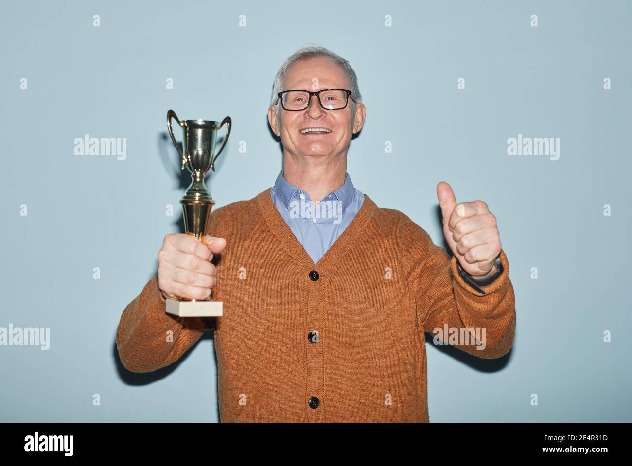 Waist up portrait of smiling senior man holding trophy and looking at camera while standing against blue background, copy space Stock Photo