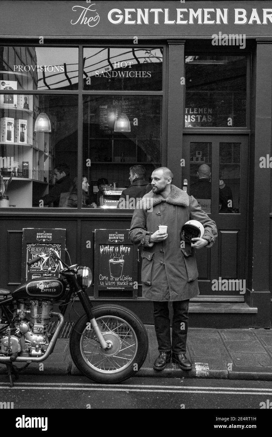 A man takes a coffee break at a cafe in London with classic motorcycle parked in front of a coffee shop Stock Photo