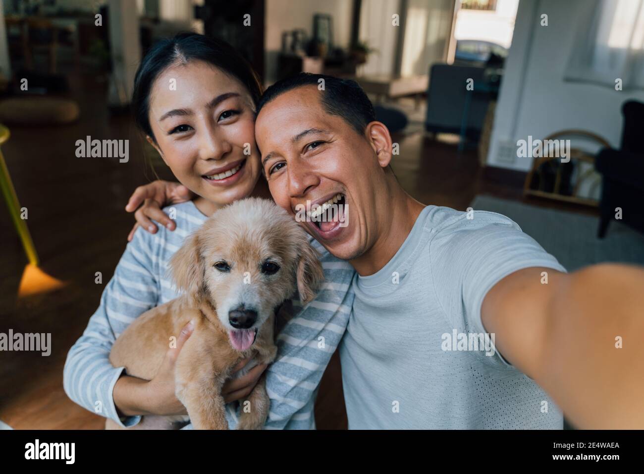 Young adult Asian couple holding a puppy taking a selfie from a phone with home interior in background. 30s mature man and woman with dog pet taking a family photo shots. - Happy group portrait Stock Photo