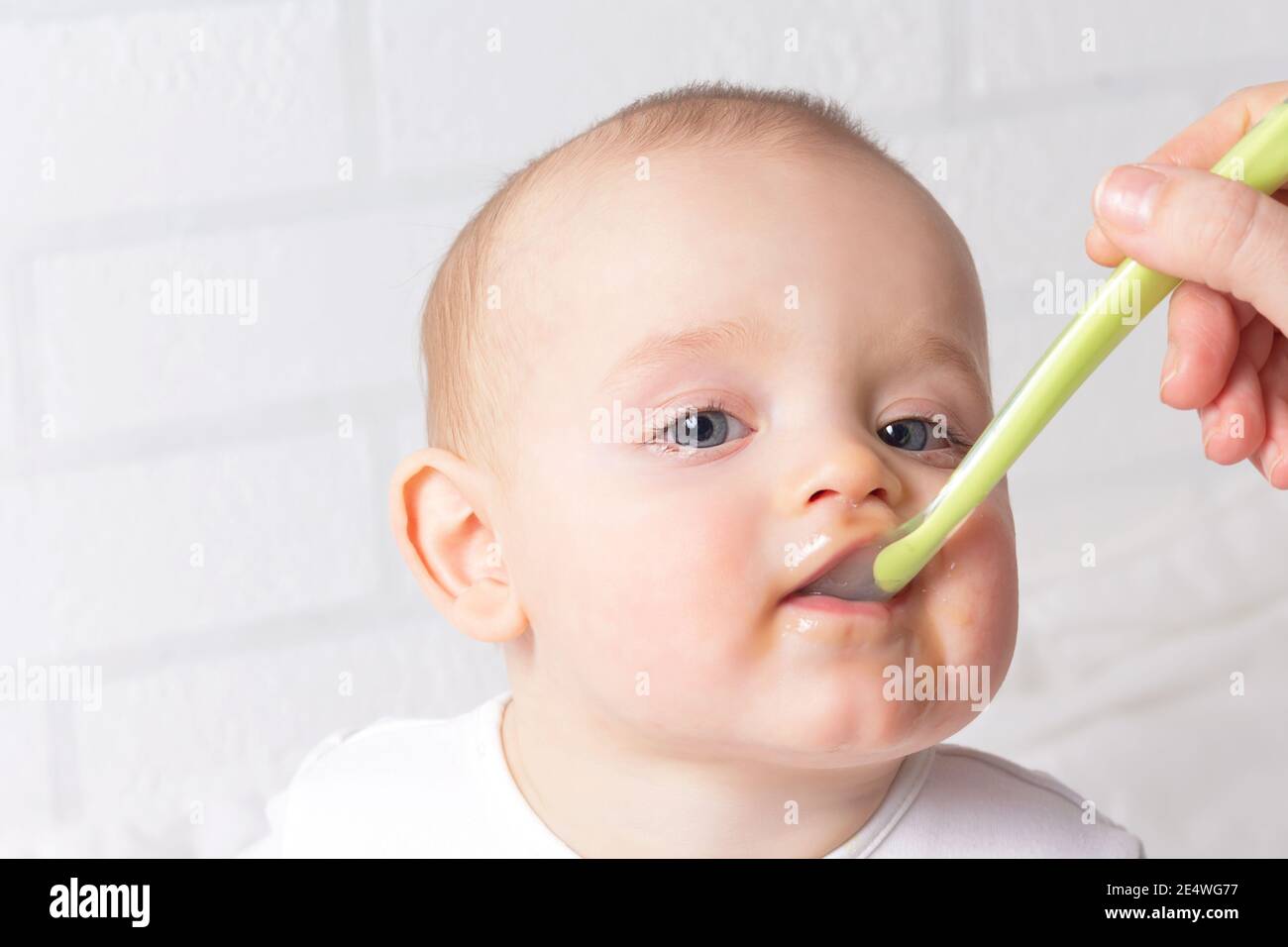 Funny caucasian baby boy getting healthy food. Fist baby food concept. Babys mouth is stained with fruit puree. Light background. Baby plastic spoon.  Stock Photo