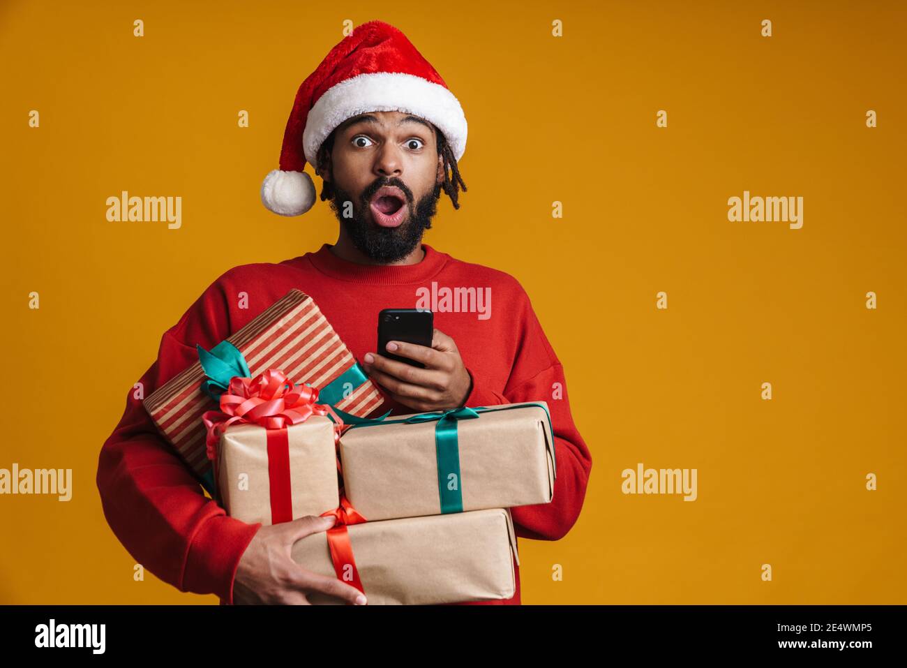 Excited young african american man in santa claus holding stack of boxes isolated on yellow background, using mobile phone Stock Photo