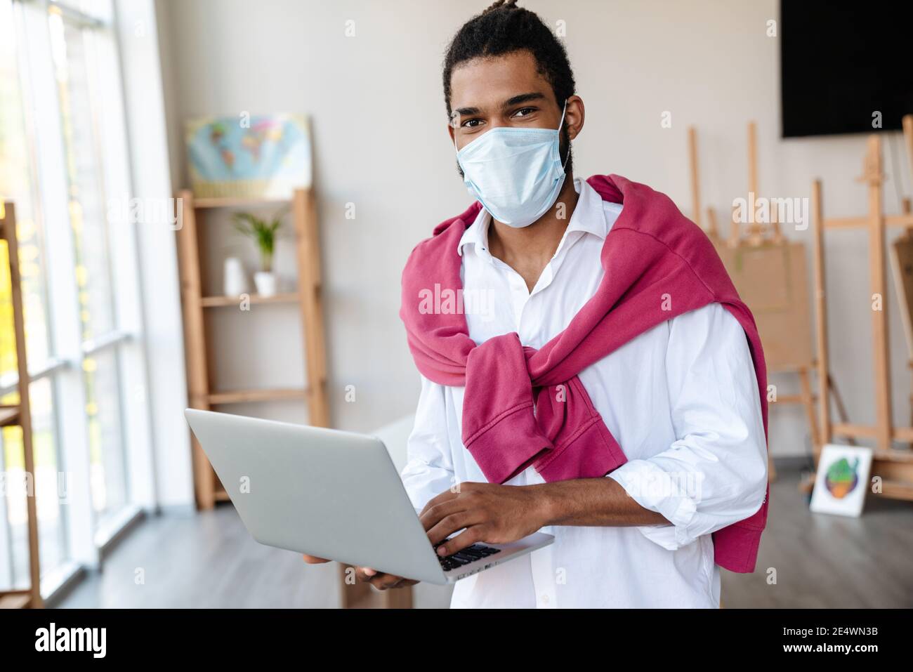 Joyful african american guy in face mask using laptop while standing at office Stock Photo