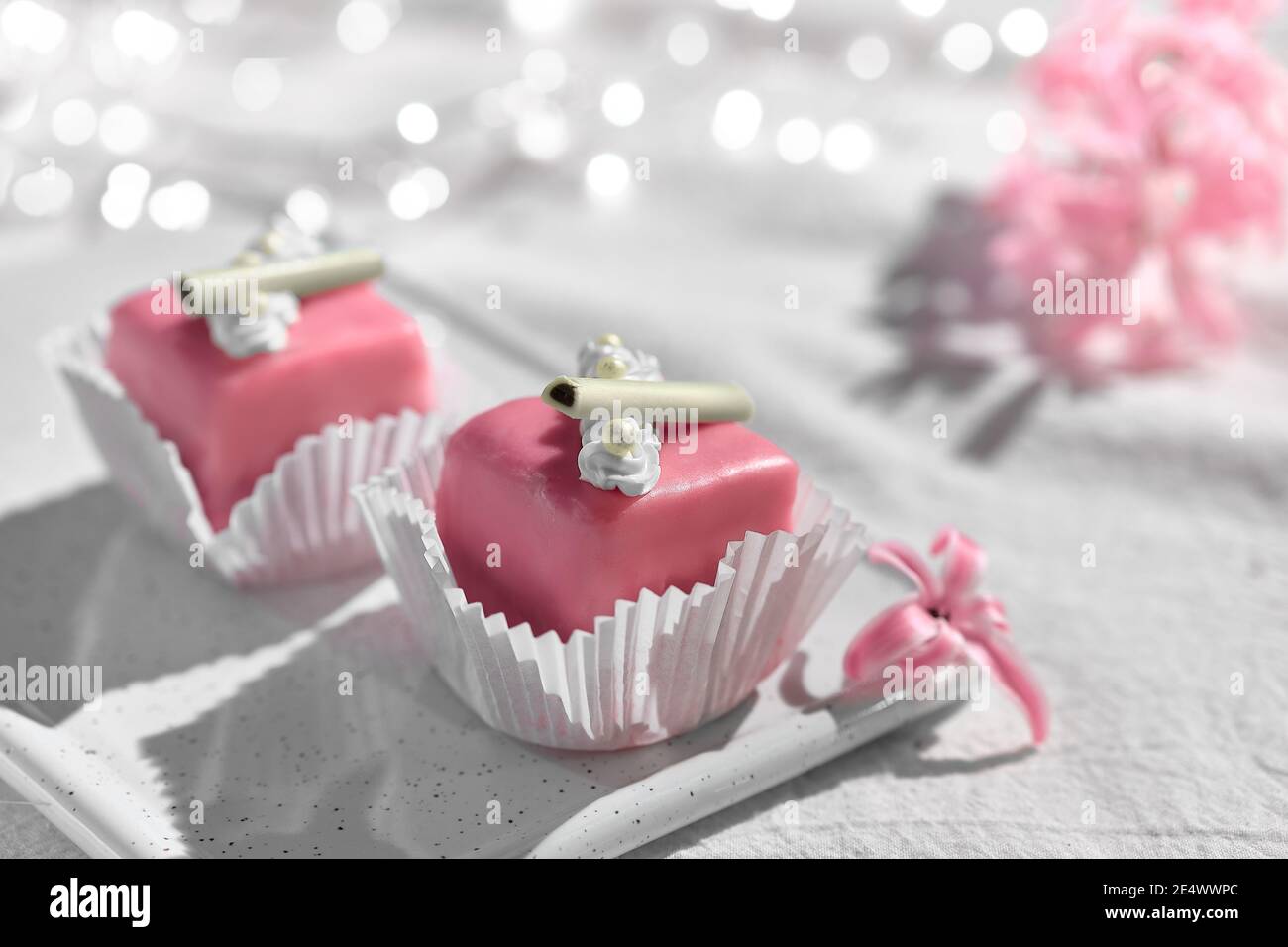 Valentine petit fours with marzipan icing. Fragrant pink hyacinth flower. Garland of lights on white textile. Romantic evening at home. Happy Stock Photo
