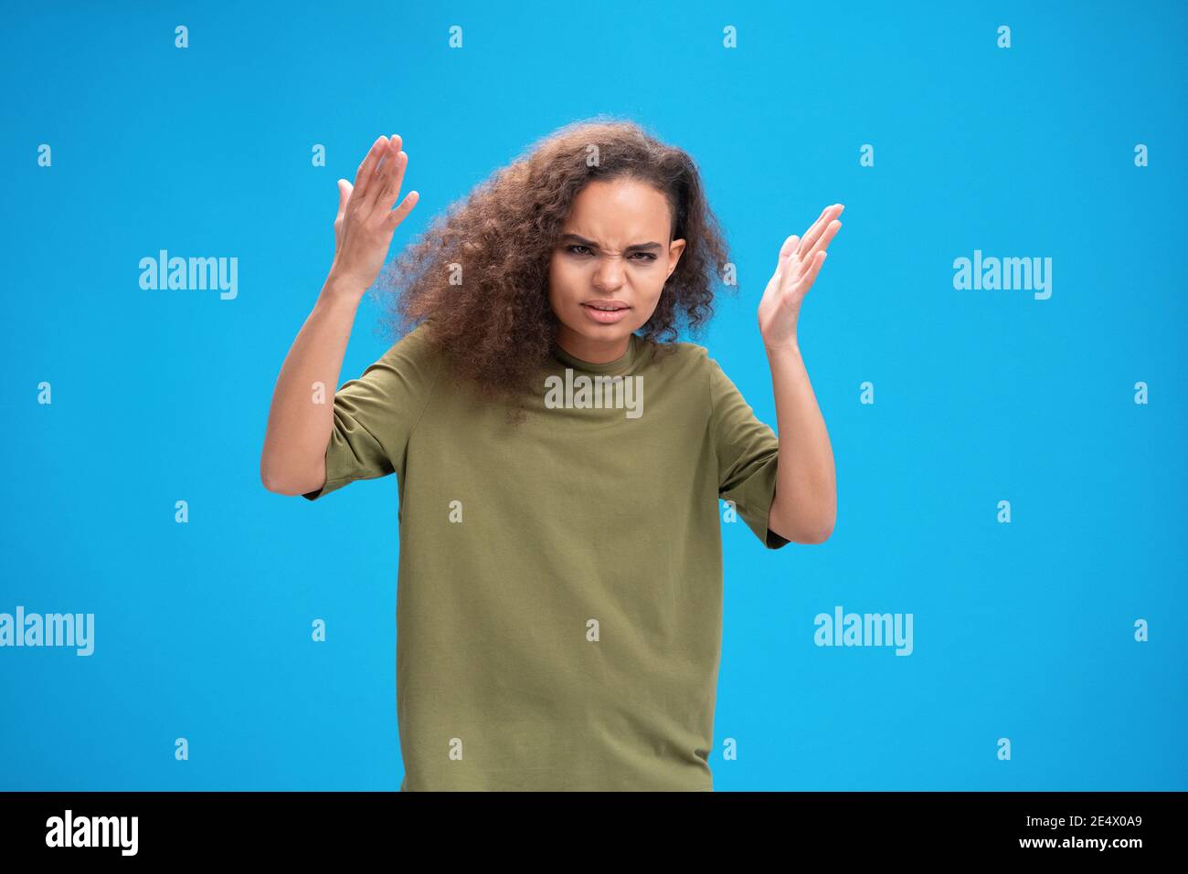 Angry african American girl lifted her hands up looking disappointedly at camera wearing olive t-shirt isolated on blue background. Human emotions Stock Photo