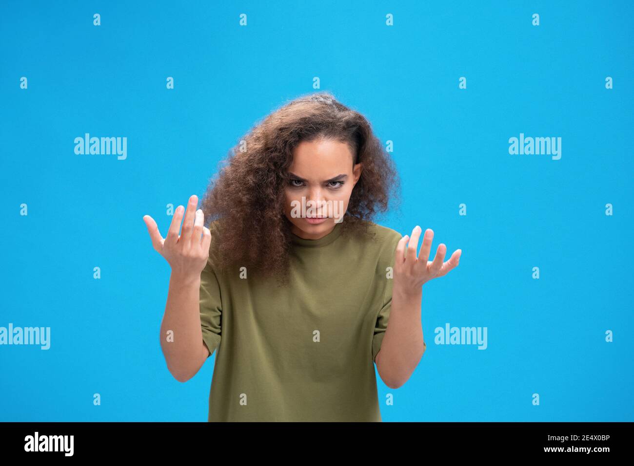 Unhappy african American millennial girl upset lifted hands up looking disappointedly at camera wearing olive t-shirt isolated on blue background Stock Photo