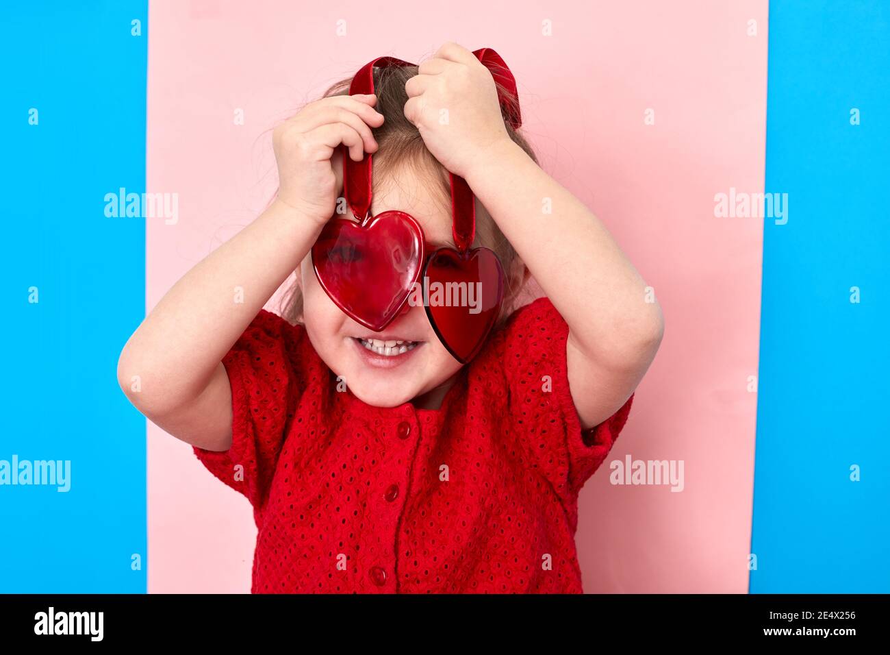 Cute delighted little girl standing in studio and looking at camera through red plastic toy hearts Stock Photo
