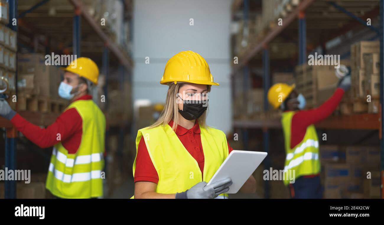 Team working in warehouse doing inventory using digital tablet and loading delivery boxes plan while wearing face mask during corona virus outbreak Stock Photo